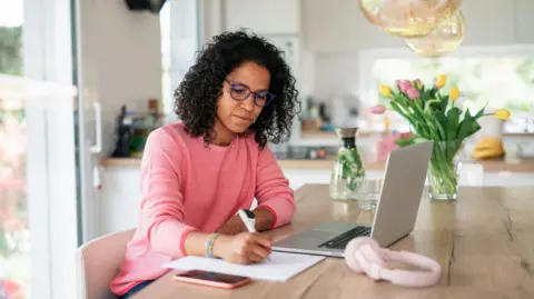 Getty Images A woman sits at her kitchen table with a laptop and pen and paper