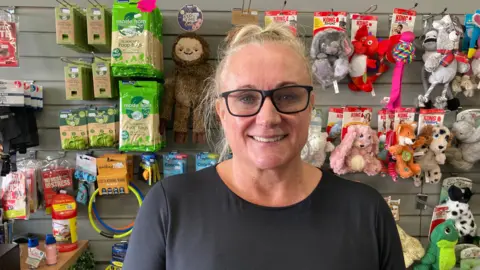 A woman with tied back blonde hair and black square glasses smiles in a shop with dog toys and treats behind her.
