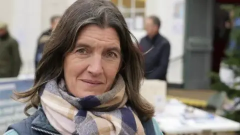 John Fairhall/BBC Rosie Pearson with mid-length brown hair looks into the camera. She is wearing a scarf with a purple, beige and pink checked pattern.