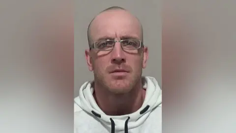 Northumbria Police Mugshot of Gavin Hall. He is wearing a white hoodie with black drawstrings, has thin stubble on his chin and top of his head and is wearing grey rectangular-framed glasses.