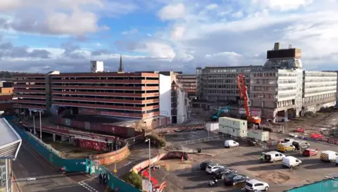 A large crushing crane and other demolition equipment are stationed next to a red-coloured multi-storey car park, which is partially demolished