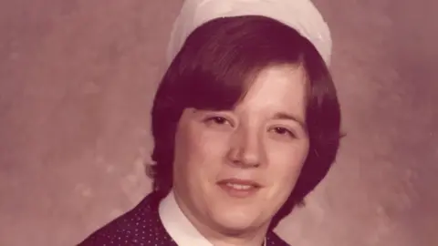 North West Anglia Foundation NHS Trust Denise, a woman, wearing a nursing white hat and a brown and white polka dot dress, smiling for camera.
