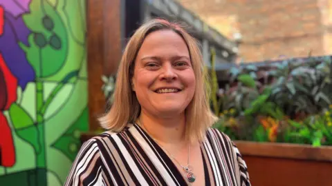 BBC/Claire Hamilton Iwonna Kossek, who is wearing a striped blouse, smiles while sitting in a restaurant. 