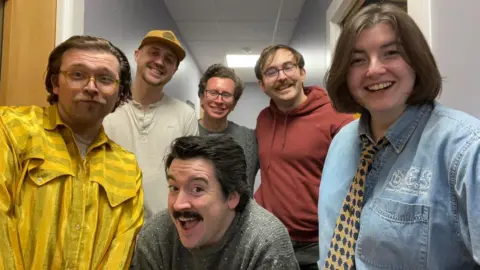 JR McCormick Six people stand together in a hallway, smiling at the camera. There are three people in the front and three people in the back. Front left is a man wearing a yellow shirt. Front middle is a man wearing a grey jumper. Front right is a woman wearing a denim shirt. Back left is a man wearing a white top and a yellow hat. Back middle is a man wearing a grey jumper. Back right is a man wearing a red hoodie. 