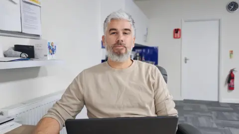 Paul Moseley/BBC Chris Gibbons sentado en su oficina, con una computadora portátil frente a él.