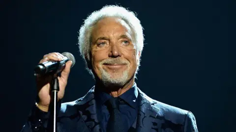 Sir Tom Jones holds a mic stand and grins