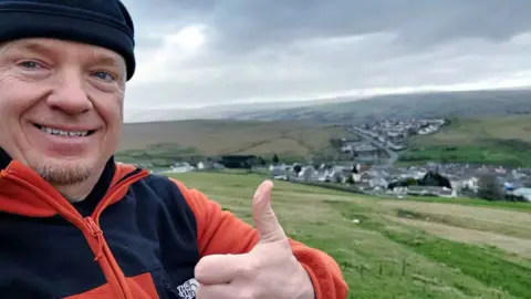 Instagram | Robert Charles Albon Robert Charles Albon looks towards the camera in the middle of a field, with green countryside behind him. He is wearing a black hat, and a black and orange jumper. He is holding his thumb up and smiling at the camera.