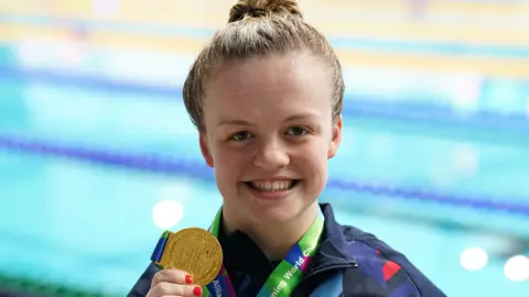 PA A smiling Maisie Summers-Newton stands beside a swimming pool and holds up her gold medal at the camera. 