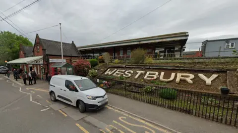 Google Westbury train station, with the word Westbury in large white letters on a hill in front of the platforms