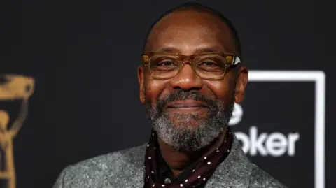 A man in a grey jacket with maroon and white spotted shirt grins with a closed mouth in front of a black wall. He is wearing light brown framed glasses and has a shaved head and greying beard.