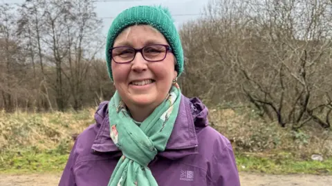 Lisa is wearing dark purple framed glasses, a green woolly hat, a scarf with prints of birds on and a purple waterproof coat. She is stood on a canal path and is smiling at the camera.