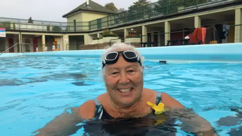 JOHN DEVINE/BBC Clare Marshall is smiling, with her neck and head poking out of the water at Peterborough Lido. She has goggles on her forehead and is wearing a blue swim suit. In the background is railings and changing rooms. 