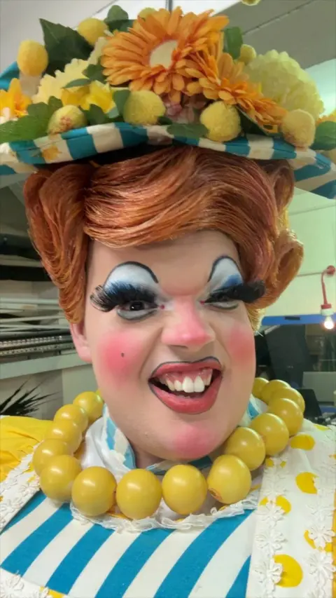 A pantomime dame wearing a big yellow necklace smiles at the camera. 