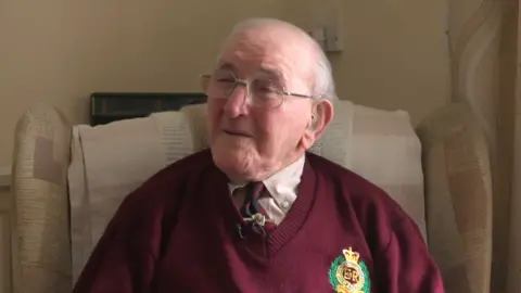 An elderly man in a maroon jumper and tie looks off the side of the camera