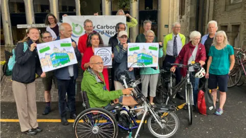 A group of people gathered outside of Oxford's county hall. One man in the foreground is on a recumbent bike and a woman holds a standard bike. Some of the people are holding signs saying "Keep Oxford's Buses Moving".