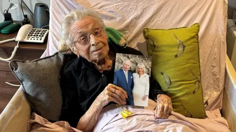 Marjorie Hodnett, a elderly lady, lies propped up in a bed, with two pillows either side of her. She has grey hair and is wearing glasses and holds up a picture of King Charles and Queen Camilla. 