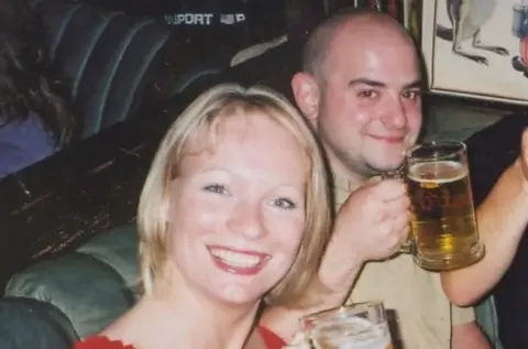 Tom and Lisa are seated together in what appears to be a pub or bar. Lisa is seated in the foreground on the left side of the photo, closest to the camera. Lisa has light-coloured, shoulder-length hair and is facing toward the camera. Tom is seated slightly behind and to the right. Tom is holding a large clear glass mug filled with an amber-coloured drink and is also facing toward the camera. 