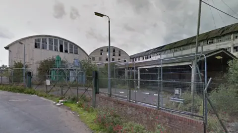 Google This picture is off the factory before the fire, and is a big white warehouse. The ceilings of the warehouse are oval shapes, and there are multiple windows at the top of the building. 