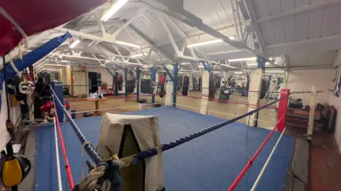 Dinesh Darbar The inside of a boxing ring, with roped sides and a blue plastic floor. 
