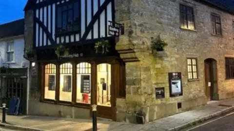 Large stone building with cash machine in its side wall, a sign on the pavement outside and it's door open.  The are several hanging baskets along its walls.