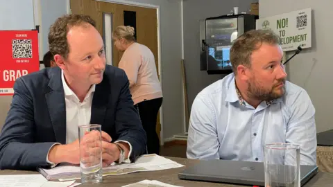 Two men sitting a desk, with a laptop, paperwork and two empty glasses. The man on the left is wearing a dark suit. The man on the right is in a light blue shirt. A placard leaning against a wall in the background reads, "No to development".