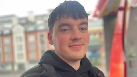 A man looks and smiles into the camera. He has mid length brown hair which has been pulled down over his forehead. He is wearing a black hoody and dark coat.