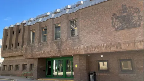A two-storey brown brick building with a green, glass-panelled door. On the front of the building is the Royal Coat of Arms. 