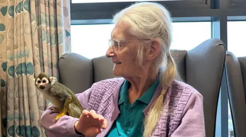 Kimberley Piper/BBC Ruth Cox has long silver hair tied up in a ponytail and is holding a squirrel monkey which is looking at the camera. She is sat in a chat and wearing a pink cardigan and green polo shirt.