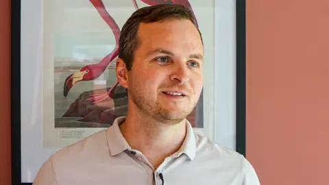Ryan Bloomfield Ryan Bloomfield stands in a room in front of a picture of a flamingo. He has brown hair, a close-cropped beard and wears a light-coloured polo shirt.