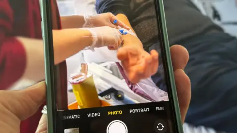 A photograph of a phone filming a patient in hospital. In the viewfinder of the phone you can see a patient's arm and a nurse giving him a drug and performing a medical procedure. 