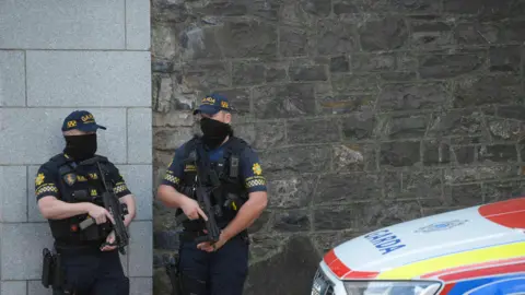 PA Media Members of the Garda Armed response unit at The Criminal Courts of Justice in Dublin. Two men stand holding large guns, their faces are covered with black coverings, they wear a blue uniform, including a hat. The building behind them i smade out of brick and they stand near a police car.