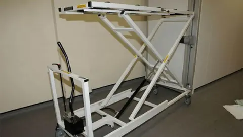 PA Media A white metal device, with a lifting bed, scissor lift and a black hydraulic pump on the bottom, in a clinical room.