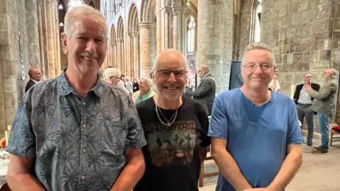 BBC/RICHARD EDWARDS Former shipbuilders Phil Moat, David McMillan and Paul Welch are standing in a line on the altar at Selby Abbey. Phil is wearing a shirt with palm trees on it, David is wearing a black T-shirt and two gold necklaces and Paul is wearing a blue T-shirt. They are all smiling.