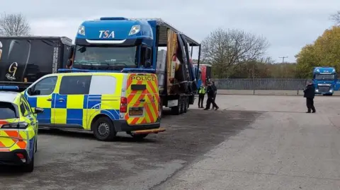 Police officers can be seen inspecting the back of a lorry. Two police vehicles are in the foreground