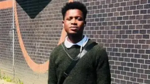 Johnson Ewere wearing a green jumper with a white polo neck underneath. He has the strap of a black bag across his body. He is standing in front of a wall with dark bricks and a large circle of orange brick.
