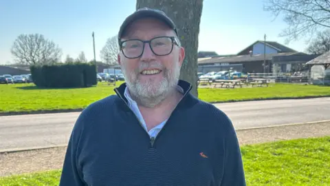 Farm Fest A man with a grey beard and in a blue fleece is by a tree smiling at the camera. A road, a building, grass and parked cars are behind him.