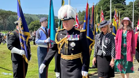 Lieutenant Governor Sir John Lorimer wearing his ceremonial uniform, which is black with gold braid, and a feathered hat. The are standards being held up alongside the walkway and his aid is walking behind him.