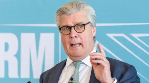 Malcolm Offord, who has grey hair and dark-rimmed glasses, speaks with his left hand raised in front of him. He is standing in front of a blue Reform-branded sign 
