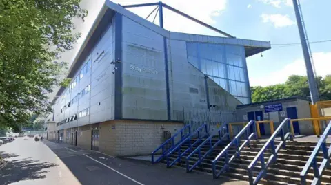 Google The sun shines above the main stand of the stadium, which is grey with blue pillars on top of light brown bricks.  In front of it are steps with thick blue bannisters leading up to the turnstiles, which have blue doors.