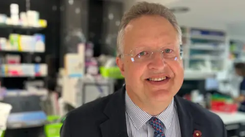 BBC A man wearing glasses a striped shirt and a jacket and a tie with a person and shelves in the background.