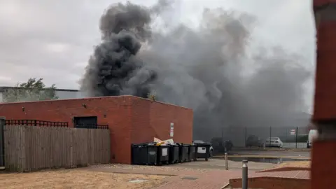Supplied A fire in a building, showing thick dark smoke coming up in the air. There are a row of black bins, cars in the distance and a building behind the smoke. 