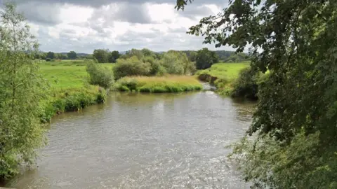 The River Lugg from Wergins Bridge near Sutton St Nicholas. The photograph shows a winding river flowing through countryside and fields and shrubs and trees overhanging the water.