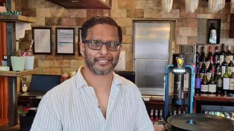 A man wearing a white shirt with black pinstripes stood in a pub.