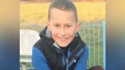 Family Handout Layton Carr. He is wearing a blue coat and is crouching down in a play area of a park. He is smiling widely at the camera.
