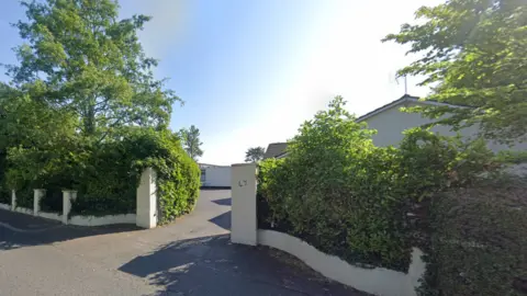 Outside street view of Somerton homes. There is a white wall with '47' on the right pillar. A number of buses and trees surround it.