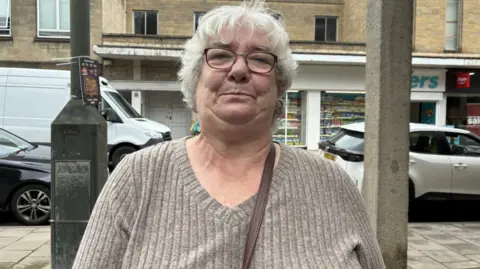 A woman in a grey jumper and black glasses with short white hair. She is stood outside on the street in front of shops, cars and a lamp post.