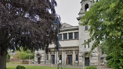 Google A church with two white stone bell towers either side of an apex roof with grey tiles. There are big trees in the foreground.
