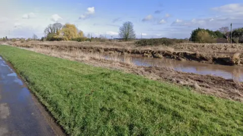 Google A river running next to a rural single-carriageway road. A short grass verge separates the road from the water.