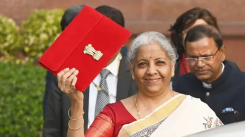 Hindustan Times via Getty Images Union Finance Minister Nirmala Sitharaman with other officials outside India's Finance Ministry before presenting last year's union budget. She's smiling at the camera in a white saree, holding a red brief case. 