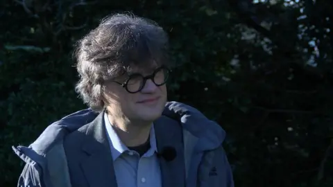  Dr Alexander Haupt looking to the right wearing black glasses, a blue coat, suit jacket and blue shirt. 